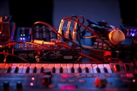 An array of musical equipment including electronic instruments, cables, and a microphone is illuminated by colorful lighting. The scene conveys an atmosphere of preparation or live performance setup.