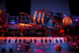An array of musical equipment including electronic instruments, cables, and a microphone is illuminated by colorful lighting. The scene conveys an atmosphere of preparation or live performance setup.