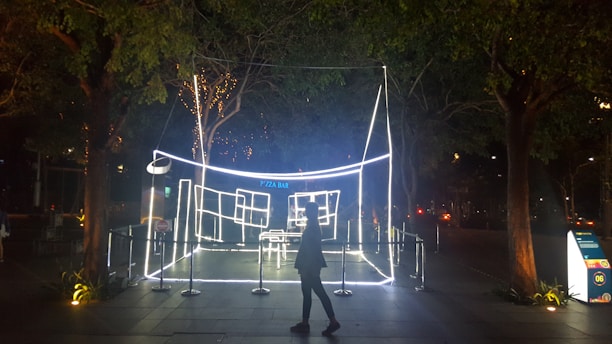 An outdoor installation featuring a structure outlined with bright white neon lights, resembling a minimalistic pizza bar. The installation is surrounded by barriers and illuminated trees, creating a striking contrast in the nighttime setting. A silhouette of a person walks past the display.