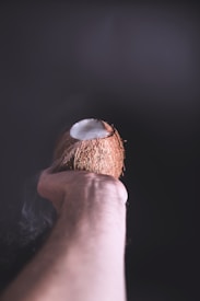 A hand is holding a halved coconut, with its textured brown shell and white flesh visible. The dark background creates a stark contrast, highlighting the coconut and the hand.