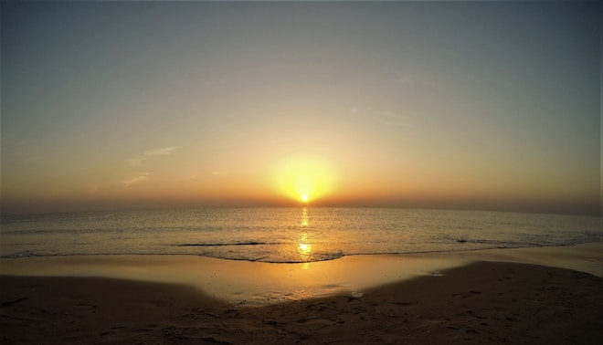 A serene beach scene at sunset with the sun setting on the horizon over a calm ocean, casting a golden glow across the water and sand.