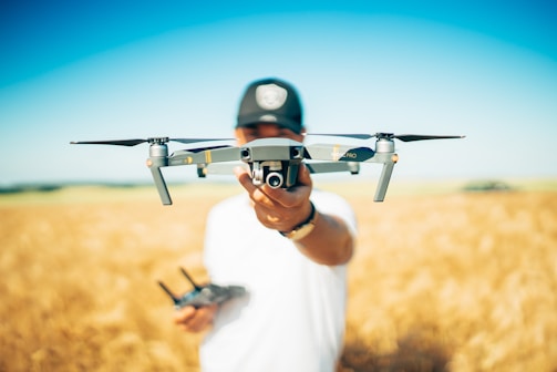 A person holds a drone prominently in the center, with a wheat field and clear blue sky in the background. The drone is in sharp focus while the person and the background are slightly blurred.