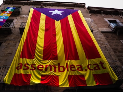 A large banner with red and yellow vertical stripes, featuring a blue triangle with a white star at the top. The text 'assemblea.cat' is visible on the yellow portion of the banner. The banner is draped over a stone building with several balconies.