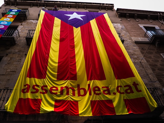 A large banner with red and yellow vertical stripes, featuring a blue triangle with a white star at the top. The text 'assemblea.cat' is visible on the yellow portion of the banner. The banner is draped over a stone building with several balconies.