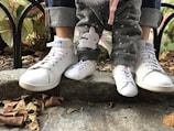 Two pairs of feet, one adult and one child, are wearing matching white sneakers. The adult is wearing cuffed jeans, and the child is in grey pants with star patterns and a rabbit print. The feet are positioned on a stone step with dry leaves scattered around, and there are some ornamental plants in the background behind a black metal fence.