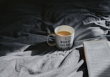 A mug with the words 'I woke up like this #tired' sits on a rumpled, dark-colored bedspread. Light casts a shadow, creating a cozy atmosphere. Next to the mug is an open book titled 'Wild at Heart' by John Eldredge. The scene suggests a quiet, leisurely moment, possibly in the morning.