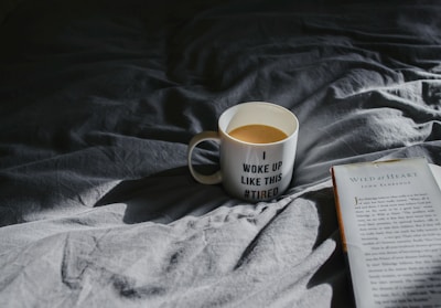 A mug with the words 'I woke up like this #tired' sits on a rumpled, dark-colored bedspread. Light casts a shadow, creating a cozy atmosphere. Next to the mug is an open book titled 'Wild at Heart' by John Eldredge. The scene suggests a quiet, leisurely moment, possibly in the morning.