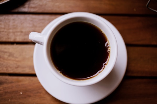 A top-down view of a cup filled with dark coffee resting on a wooden table. The white cup and saucer contrast with the rich, dark color of the coffee. The table has a natural wood grain that adds to the warmth of the composition.