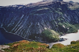 A breathtaking, vast mountain landscape featuring steep rocky cliffs and verdant patches of greenery extending down to a dark blue fjord. The scene conveys a sense of untouched natural beauty and grandeur.