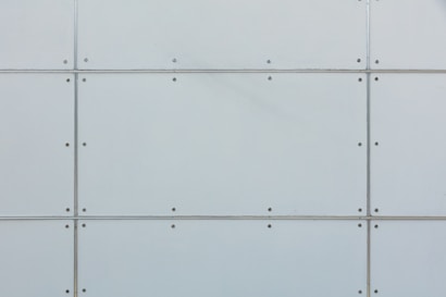 A close-up view of a metal panel wall. The structure is geometric, consisting of rectangular sections joined together by visible screws. The surface is a light gray color and has a smooth texture.