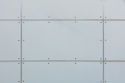 A close-up view of a metal panel wall. The structure is geometric, consisting of rectangular sections joined together by visible screws. The surface is a light gray color and has a smooth texture.