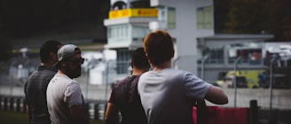 Four individuals stand near a racetrack or motorsport area, with attention focused on the track or events happening beyond. They are casually dressed, and some appear to be talking among themselves. In the background, there are buildings and barriers, typical of a racing venue.