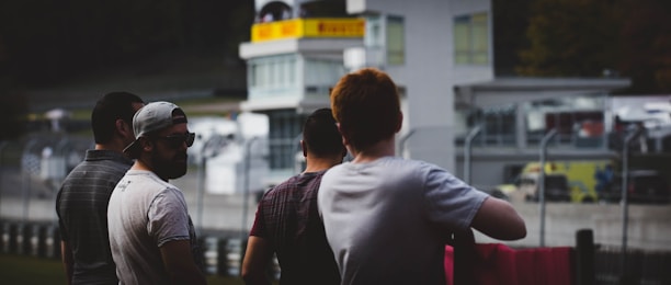 Four individuals stand near a racetrack or motorsport area, with attention focused on the track or events happening beyond. They are casually dressed, and some appear to be talking among themselves. In the background, there are buildings and barriers, typical of a racing venue.