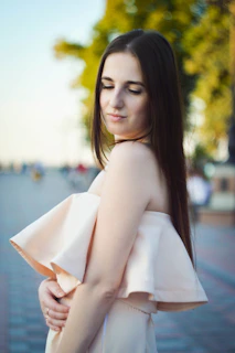 Model wearing a flowing beige dress standing in soft sunlight with a serene expression.