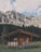 A rustic wooden cabin is nestled in a lush green meadow, set against the backdrop of towering, rugged mountains. The sky is partly cloudy, casting soft shadows over the landscape. Dense forest lines the base of the mountains, framing the secluded cabin.