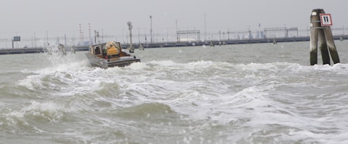 A small boat navigating through choppy waters with visible waves and splashes, moving towards a distant industrial background with structures and poles. On the right is a marker or buoy labeled with the number 11.
