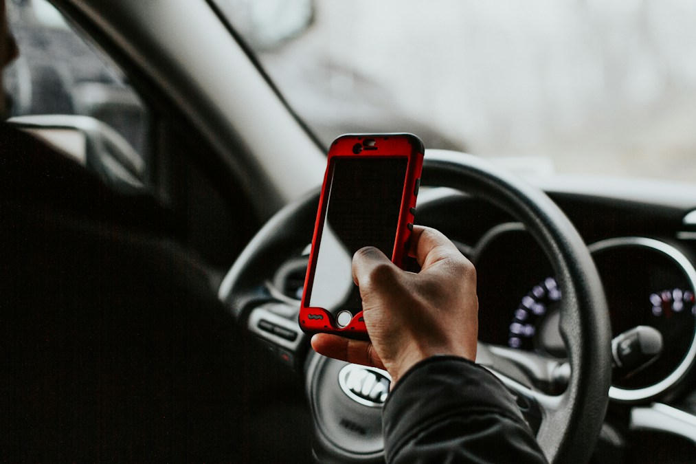 Main tenant un smartphone à coque rouge devant le volant d’une voiture, le geste que les futures caméras ANPR doivent repérer