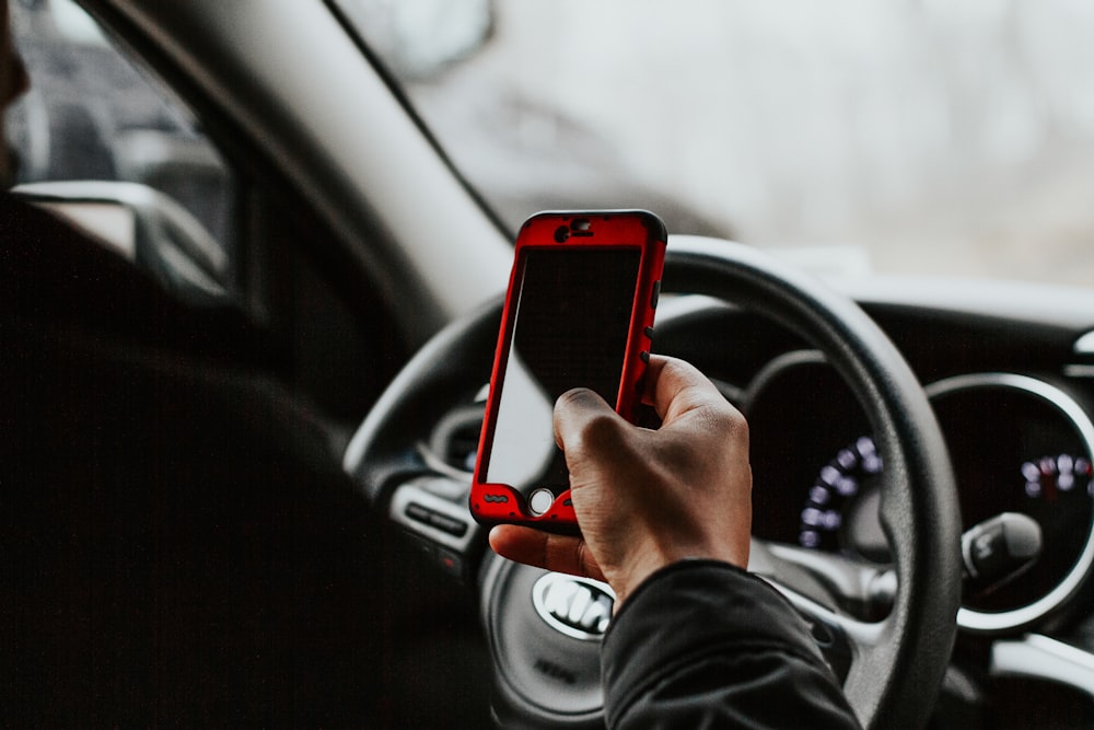 Person mit rotem Smartphone sitzt vor dem Lenkrad des Fahrzeugs