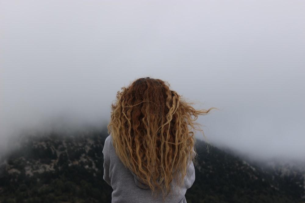mulher vestindo camisa cinza de frente para a foto da montanha verde coberta de nevoeiro