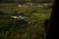 A serene rural landscape featuring a large house with a blue swimming pool, surrounded by dense greenery and trees. In the background, there are well-manicured lawns, a golf course, and a small lake with buildings near it. A road winds through the area, leading to the house and beyond.