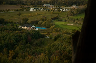 A serene rural landscape featuring a large house with a blue swimming pool, surrounded by dense greenery and trees. In the background, there are well-manicured lawns, a golf course, and a small lake with buildings near it. A road winds through the area, leading to the house and beyond.