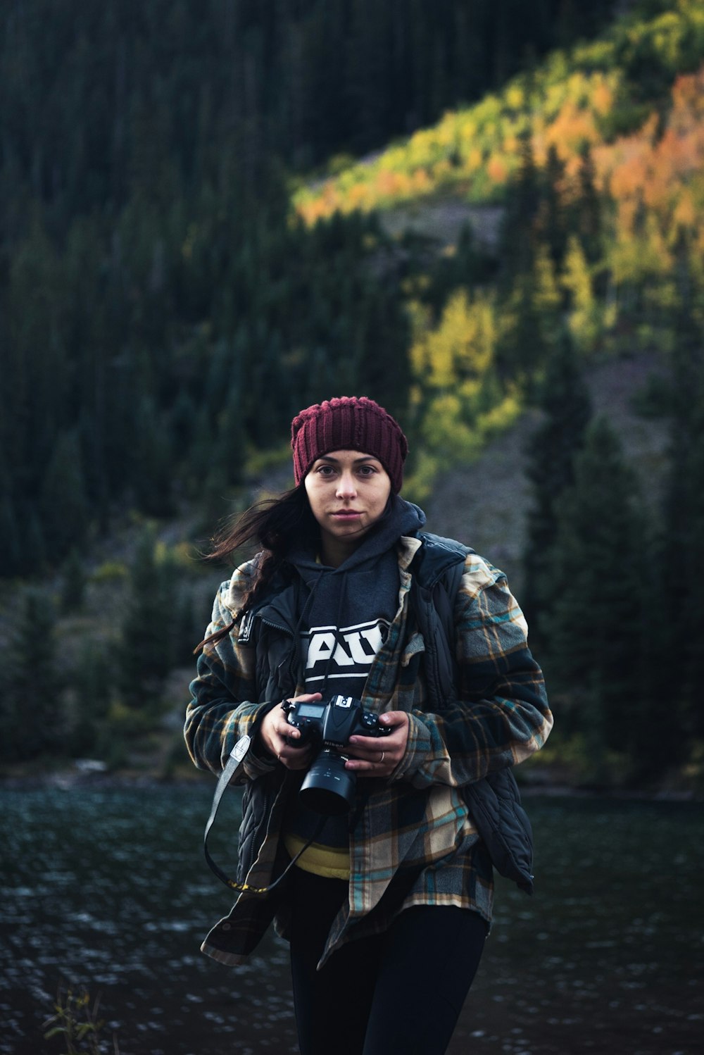 women wearing jacket and red knit cap holding DSLR camera walking near trees