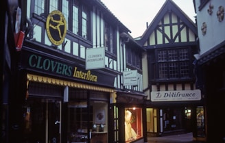 The scene depicts a quaint, narrow street lined with half-timbered buildings featuring Tudor-style architecture. The storefronts display signs for 'Clovers Interflora' and a delicatessen named 'Délifrance.' The alley has an old-world charm, with window displays showcasing various goods.