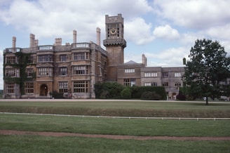 A large, historic stone building with multiple chimneys and a prominent clock tower, partially covered in ivy, is set amidst well-maintained lawns and lush greenery. The architecture suggests a heritage or academic institution, likely dating back several centuries.