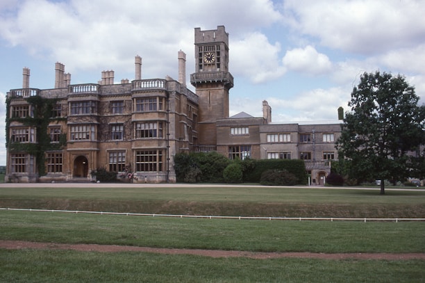 A large, historic stone building with multiple chimneys and a prominent clock tower, partially covered in ivy, is set amidst well-maintained lawns and lush greenery. The architecture suggests a heritage or academic institution, likely dating back several centuries.