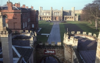 A historic castle with large stone walls and battlements, overlooking a wide grassy courtyard. The entrance is marked by a sign reading 'Welcome to Lincoln Castle'. Surrounding the courtyard are red brick buildings and trees, with a prominent stone building featuring arched windows and towers in the background.