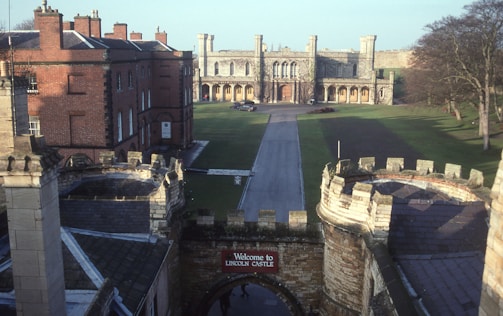 A historic castle with large stone walls and battlements, overlooking a wide grassy courtyard. The entrance is marked by a sign reading 'Welcome to Lincoln Castle'. Surrounding the courtyard are red brick buildings and trees, with a prominent stone building featuring arched windows and towers in the background.
