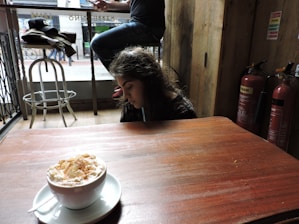 A young person sits at a table inside a cafe, looking down. A frothy beverage in a white cup is placed on the table in the foreground. In the background, a jacket and cap rest on a high chair while two fire extinguishers are mounted on a wooden wall. A street scene with a person's legs and a bicycle is visible through the window.