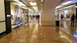 A spacious shopping mall corridor with reflective marble flooring. Storefronts line both sides, displaying mannequins dressed in various outfits. Shoppers are casually walking, adding to the leisurely atmosphere. The lighting is bright and evenly distributed, enhancing the polished look of the interior.