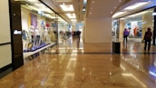 A spacious shopping mall corridor with reflective marble flooring. Storefronts line both sides, displaying mannequins dressed in various outfits. Shoppers are casually walking, adding to the leisurely atmosphere. The lighting is bright and evenly distributed, enhancing the polished look of the interior.