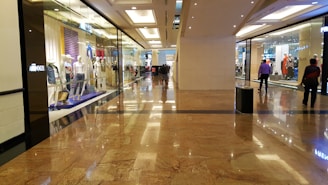 A spacious shopping mall corridor with reflective marble flooring. Storefronts line both sides, displaying mannequins dressed in various outfits. Shoppers are casually walking, adding to the leisurely atmosphere. The lighting is bright and evenly distributed, enhancing the polished look of the interior.