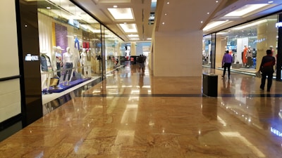A spacious shopping mall corridor with reflective marble flooring. Storefronts line both sides, displaying mannequins dressed in various outfits. Shoppers are casually walking, adding to the leisurely atmosphere. The lighting is bright and evenly distributed, enhancing the polished look of the interior.