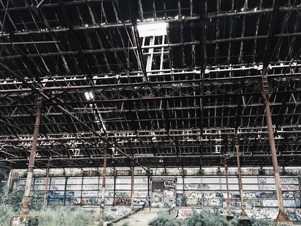 An abandoned industrial warehouse with a partially collapsed roof, featuring exposed metal beams and rusty columns. The walls are covered in colorful graffiti, and overgrown vegetation is visible on the ground.