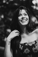 A black and white photograph of a smiling woman with long hair, wearing a floral patterned top. She has a tattoo of an anchor on her forearm and appears to be in a natural setting with blurred foliage in the background.