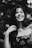A black and white photograph of a smiling woman with long hair, wearing a floral patterned top. She has a tattoo of an anchor on her forearm and appears to be in a natural setting with blurred foliage in the background.