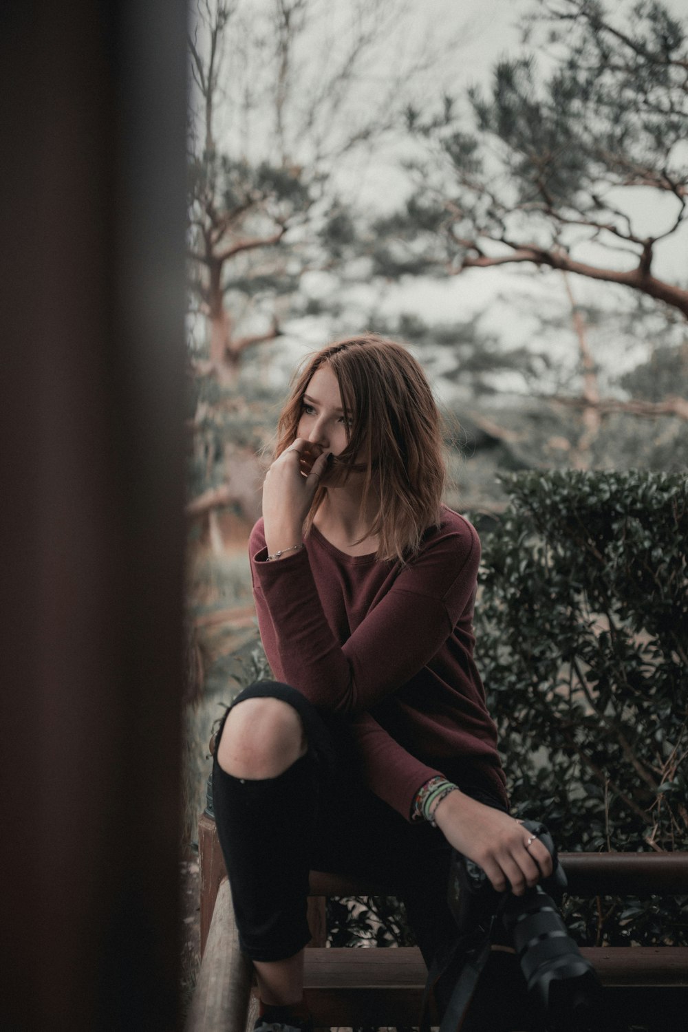 wide-aperture photography of woman sitting on beige wooden frame near green shrub under gray clouded sky during daytime