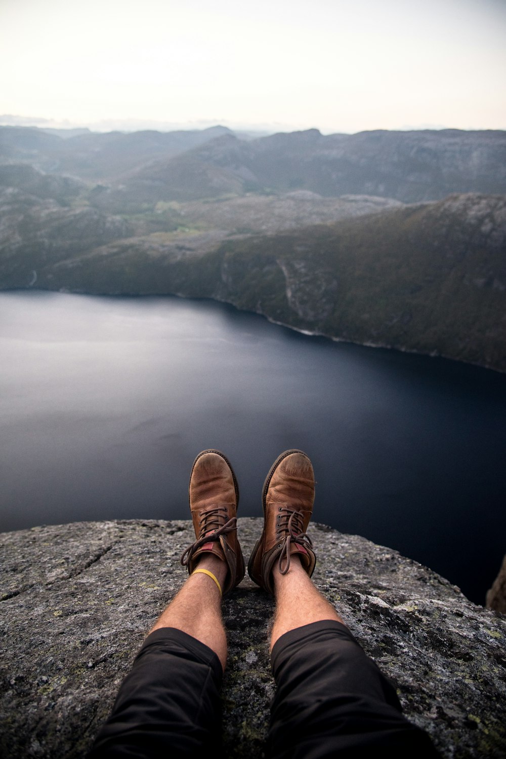 person wearing brown boots sitting on cliff in front of grey calm body of water and rock formations during daytime