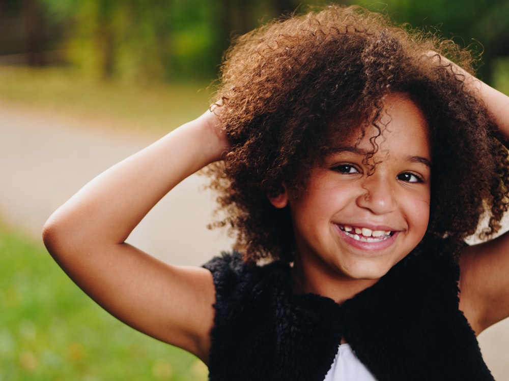 girl wearing black vest raising two hands near green grass field during daytime