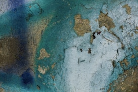 A textured wall surface with peeling paint, displaying an array of colors including shades of blue and turquoise with exposed patches of brown and beige. The pattern created by the cracks and peels gives an abstract, rustic appearance.