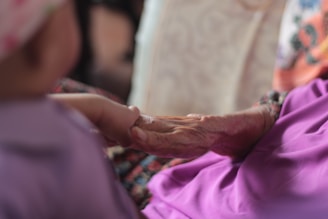 An elderly hand gently holding the hand of a younger person. The focus is on the hands, conveying a sense of tenderness and connection. The background is softly blurred, emphasizing the moment of intergenerational touch.