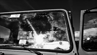 A monochrome photo captures the windshield of a vehicle with clouds and sky reflected on its surface. Inside, a stuffed toy with a snowflake design sits on the dashboard. A camera and some electronic gadgets are visible through the glass.