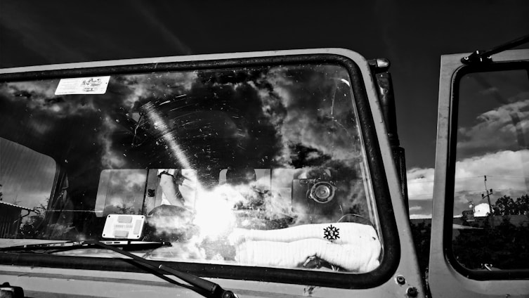 A monochrome photo captures the windshield of a vehicle with clouds and sky reflected on its surface. Inside, a stuffed toy with a snowflake design sits on the dashboard. A camera and some electronic gadgets are visible through the glass.