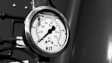 A close-up view of a pressure gauge with a black dial and white background, displaying measurements in psi and bar. The gauge is marked 'KIT' at the bottom, and there is a small fly resting on the glass surface.