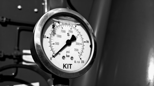 A close-up view of a pressure gauge with a black dial and white background, displaying measurements in psi and bar. The gauge is marked 'KIT' at the bottom, and there is a small fly resting on the glass surface.