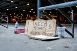 A cardboard sign with handwritten text that reads 'Need Some $ Help Used Books' is propped against a metal railing. Nearby, there is a plastic cup labeled 'Help' with a dollar sign, a bottle of water, and a stack of books. The scene is set under an overpass with blurred lights and parked cars in the background.