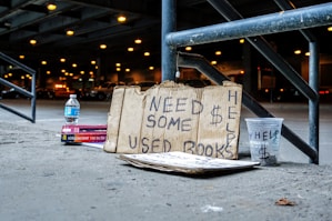 A cardboard sign with handwritten text that reads 'Need Some $ Help Used Books' is propped against a metal railing. Nearby, there is a plastic cup labeled 'Help' with a dollar sign, a bottle of water, and a stack of books. The scene is set under an overpass with blurred lights and parked cars in the background.